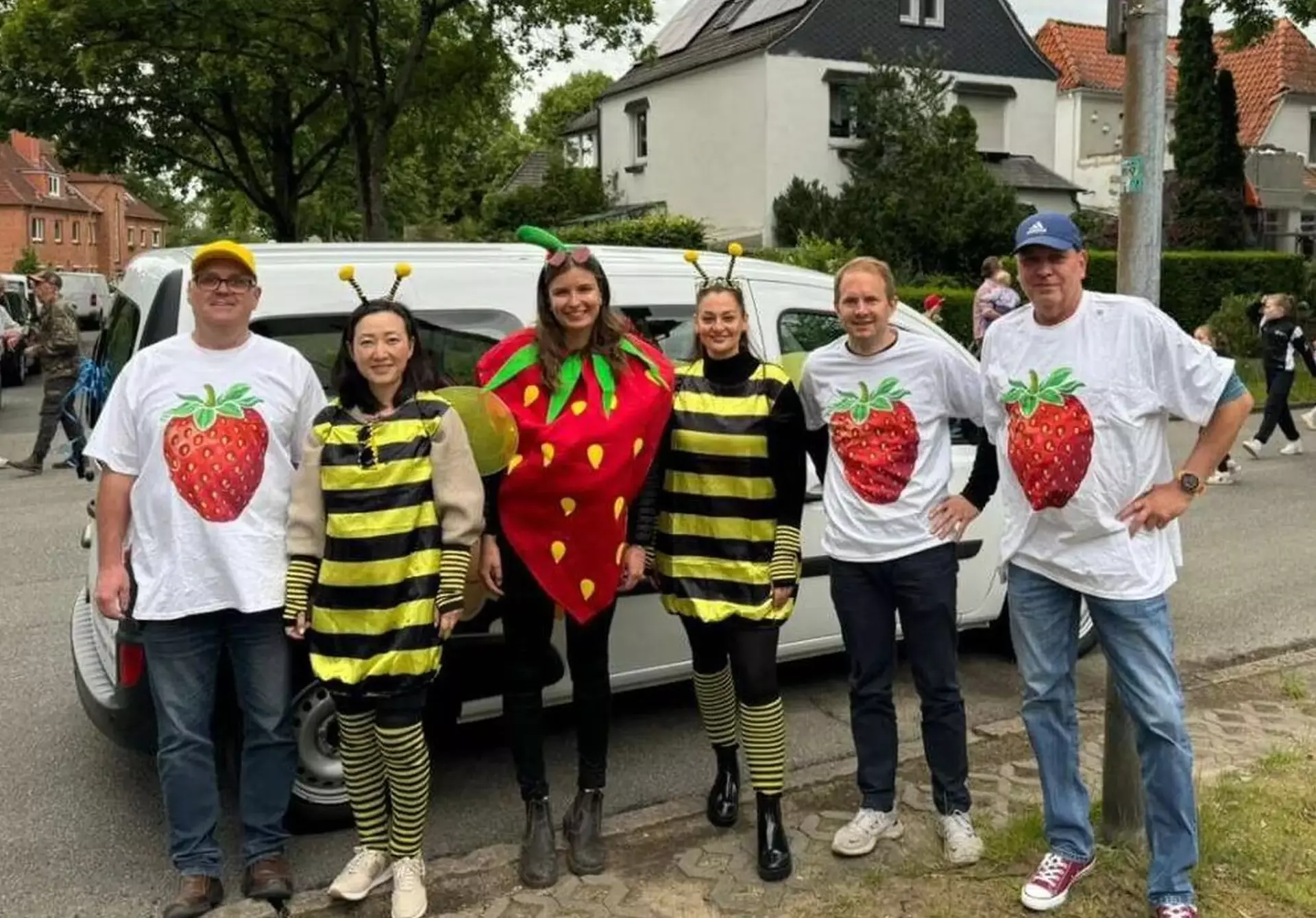 Man sieht drei Männer mit identischen T-Shirts mit großem Erdbeerdruck, zwei Frauen in Bienenkostümen und eine Frau im Erdbeerkostüm. Alle stehen als Gruppe vor einem großen Fahrzeug. Im Hintergrund sieht man einen Festumzug. 