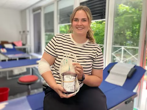 Eine Mitarbeiterin sitzt auf einer Liege in einem hellen Raum und hält einen gefüllten Blutbeutel in der Hand; am Arm ist ein Verband nach der Blutspende zu sehen.