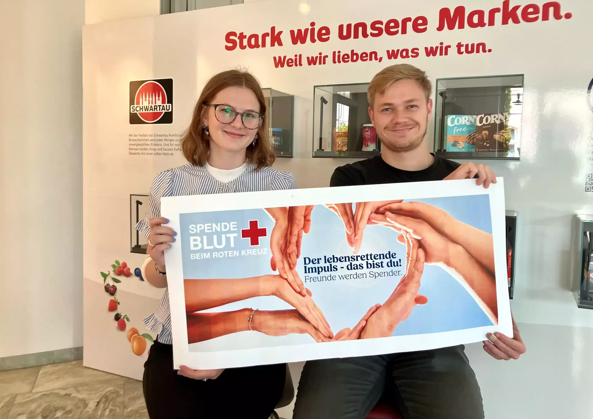 Unsere Azubis Emily und Magus aus dem 2. Lehrjahr der Industriekaufleute sitzen vor einer Wand mit Markenlogos und halten ein Banner mit der Aufschrift: ‚SPENDE BLUT BEIM ROTEN KREUZ – Der lebensrettende Impuls – das bist du! Freunde werden Spender.‘ Im Hintergrund sind Produktdisplays und der Schriftzug ‚Stark wie unsere Marken. Weil wir lieben, was wir tun.‘ zu sehen.