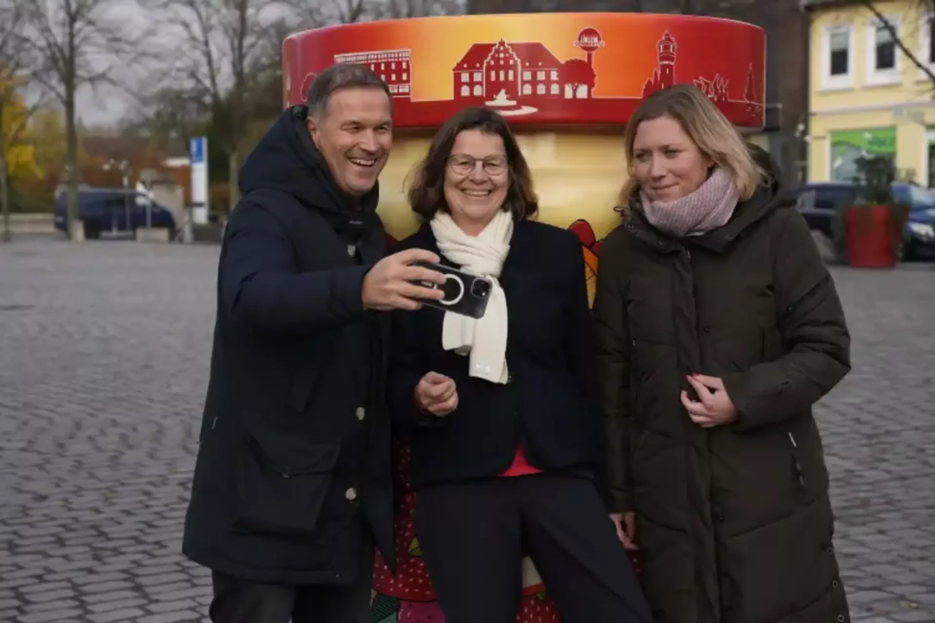 Links steht ein Mann, daneben zwei Frauen, die gemeinsam vor dem Fotopoint "Marmeladenstadt" das erste Selfie nach der Eröffnung machen.