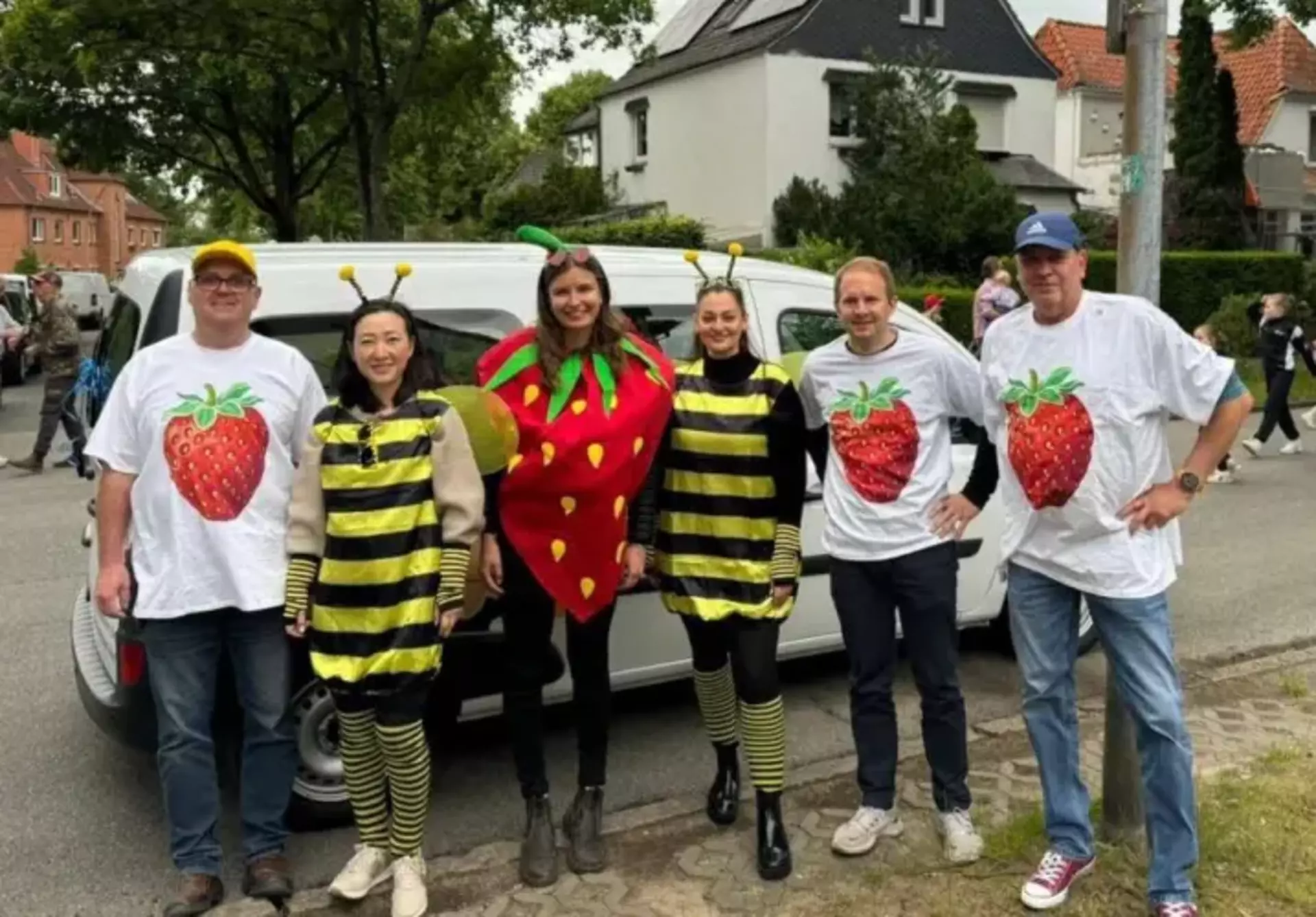 Man sieht drei Männer mit identischen T-Shirts mit großem Erdbeerdruck, zwei Frauen in Bienenkostümen und eine Frau im Erdbeerkostüm. Alle stehen als Gruppe vor einem großen Fahrzeug. Im Hintergrund sieht man einen Festumzug. 