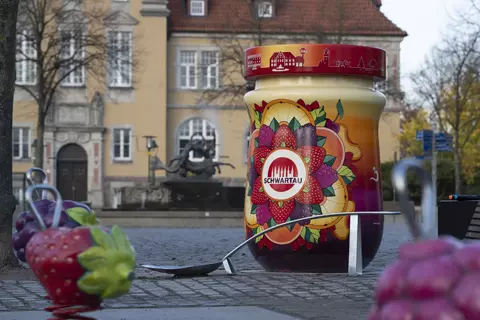 Überdimensioniertes, bunt gestaltetes Schwartau-Marmeladenglas steht auf einem öffentlichen Platz vor einem historischen Gebäude. Um das Glas herum befinden sich große, dekorative Früchte und ein übergroßer Metalllöffel als Teil einer Installation.