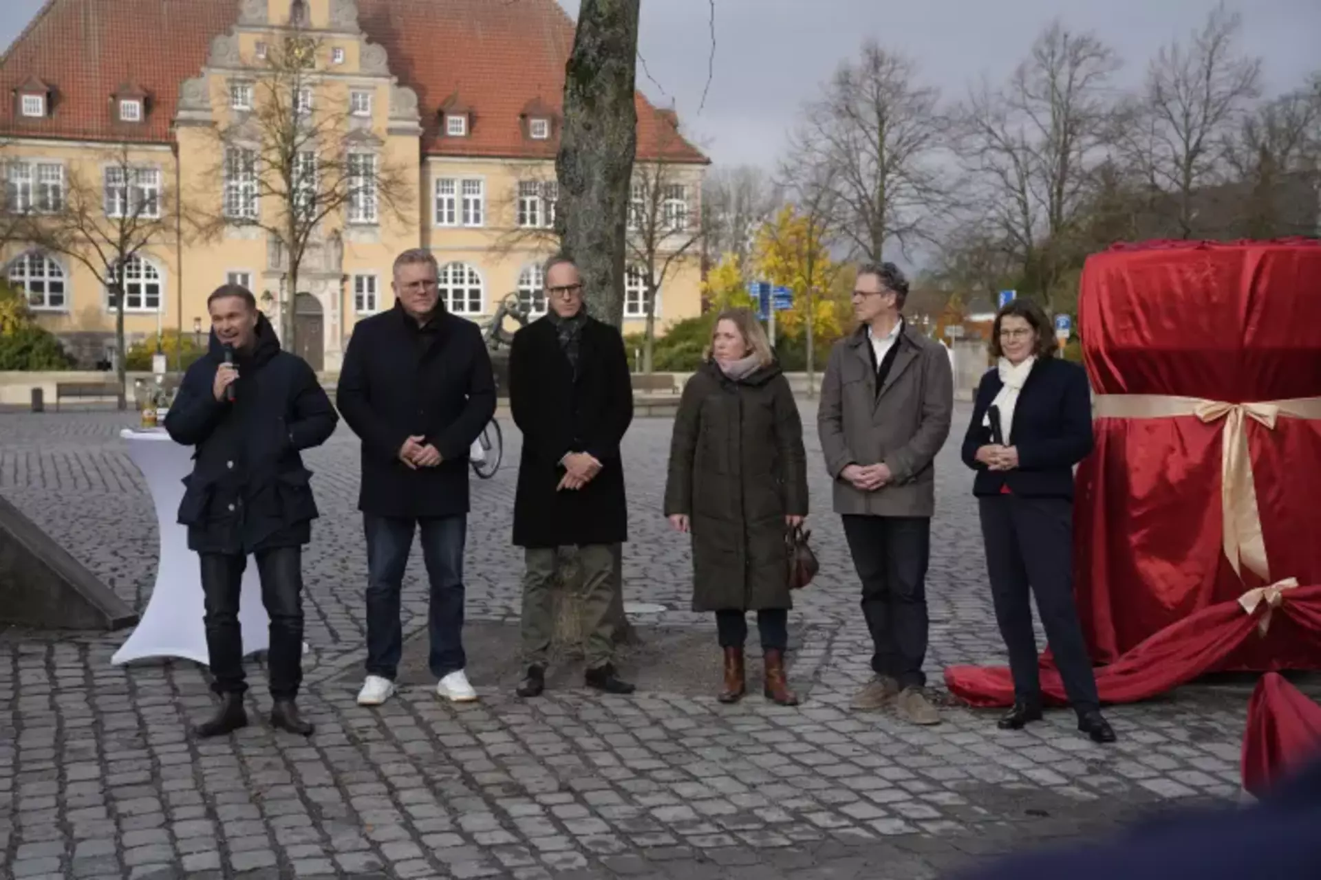 Auf dem Bild stehen sechs Personen, vier Herren von den Schwartauer Werken und zwei Damen der Stadt Bad Schwartau neben einem verfüllten Objekt in rot. Der Mann ganz links hält mit einem Mikrofon eine Ansprache.