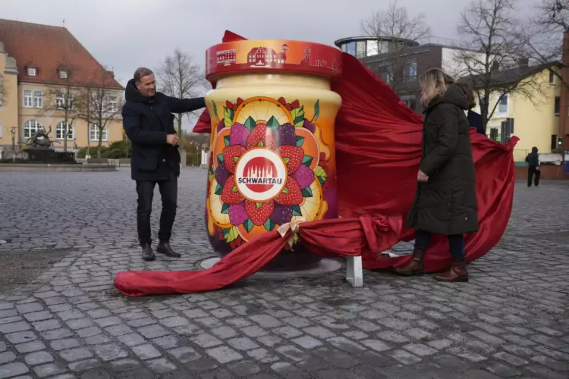 Auf dem Bild sieht man neben zwei Personen das enthüllte Kunstwerk in Form eines menschengroßen Schwartau-Marmeladenglases. Im Hintergrund sieht man noch den roten Stoff, der das Kunstwerk verdeckt hat.