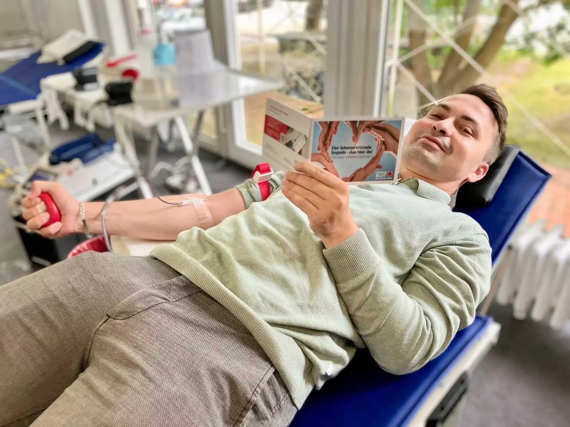 Hannes aus dem Marketing spendet Blut auf einer medizinischen Liege, hält ein Informationsheft und drückt mit der linken Hand einen roten Stressball – im Hintergrund medizinisches Equipment und große Fenster mit Tageslicht.