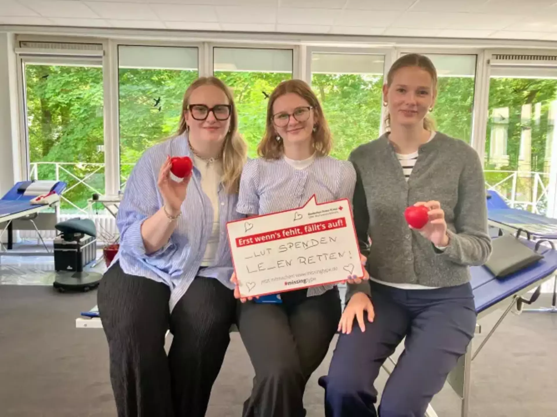 Drei Auszubildende Carlotta, Emily und Finja der Schwartauer Werke halten ein Schild mit der Aufschrift „Blut spenden – Leben retten“ und rote Herz-Stressbälle in der Hand.