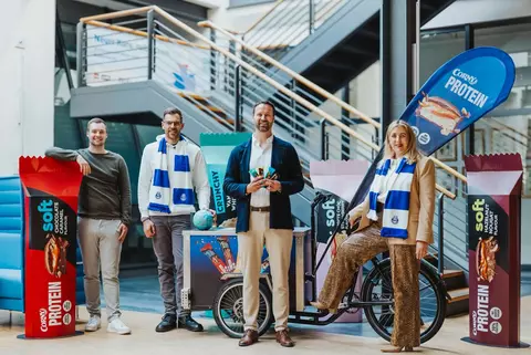 Auf dem Bild sieht man vier Personen: v. l. n. r: Lasse Ohde (VfL), Christian Kamphoven (Schwartau), Daniel Pankofer (VfL) & Vanessa Krabiell (Schwartau), die beiden Personen von Schwartau haben einen VfL-Schal um. Die beiden Personen vom VfL haben Corny-Proteinriegel in der Hand.Im Hintergrund sieht man große Corny Protein-Riegel und ein Corny Protein Lastenrad.