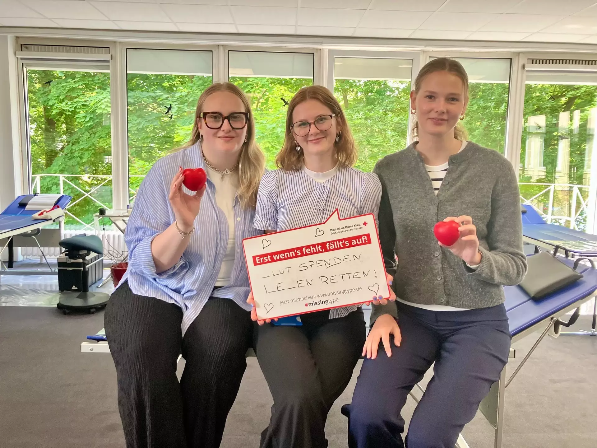Drei Auszubildende Carlotta, Emily und Finja der Schwartauer Werke halten ein Schild mit der Aufschrift „Blut spenden – Leben retten“ und rote Herz-Stressbälle in der Hand.