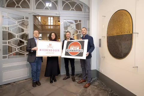 Vier Personen stehen in einem historischen Innenraum und präsentieren Schilder der Marken Niederegger Lübeck und Schwartau vor einer Eingangstür.