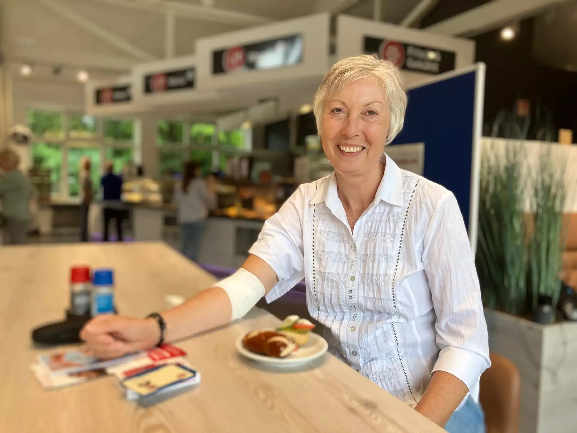 Petra aus dem Vertrieb sitzt nach ihrer Blutspende und einem kleinen Verband am rechten Arm in unserem Betriebsrestaurant an einem Tisch mit einem Teller Essen, umgeben von Salz- und Pfefferstreuern, Servietten und Informationsmaterialien – im Hintergrund weitere Personen und Essensausgabe.