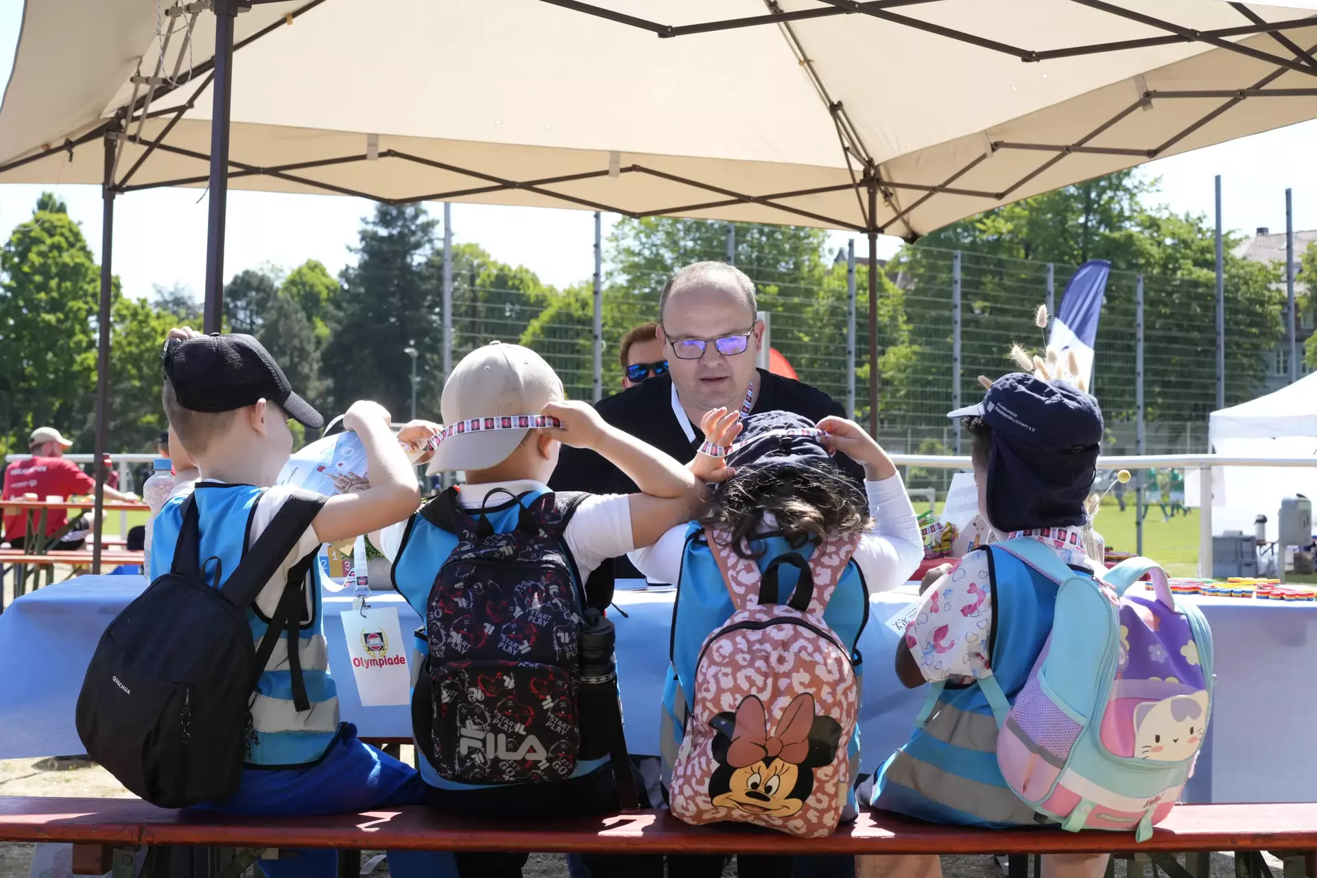 Das Foto zeigt vier Kinder, die auf einer Bank sitzen und von hinten zu sehen sind. Sie tragen Rucksäcke und Hüte oder Mützen. Die Kinder sitzen unter einem großen Sonnenschirm an einem Tisch, an dem ein Mitarbeiter der Schwartauer Werke die Nasenolympia durchführt. 