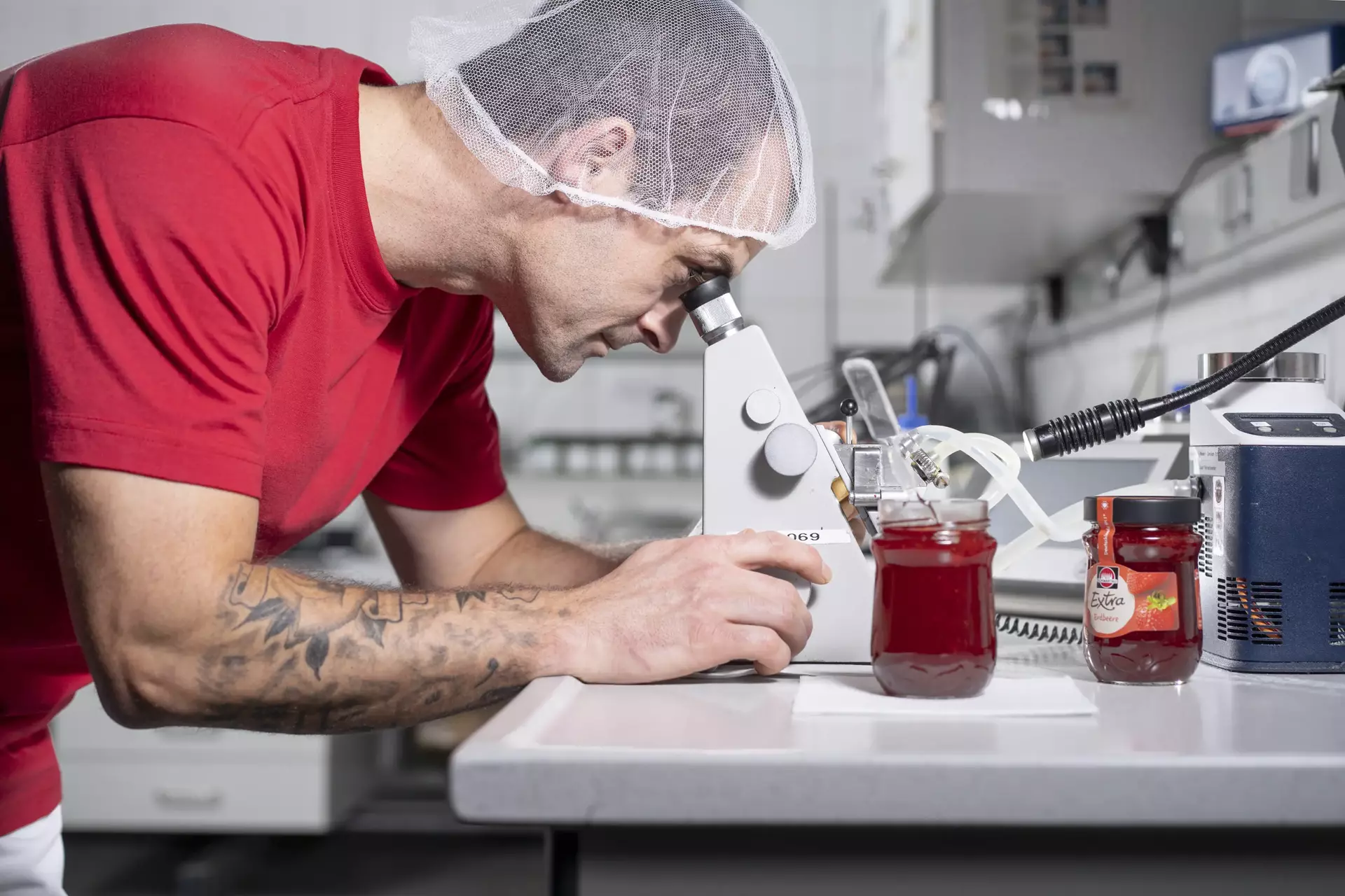 Ein Mitarbeiter an einem Mikrokop bei der Qualitätskontrolle, daneben steht ein Glas Erdbeerkonfitüre