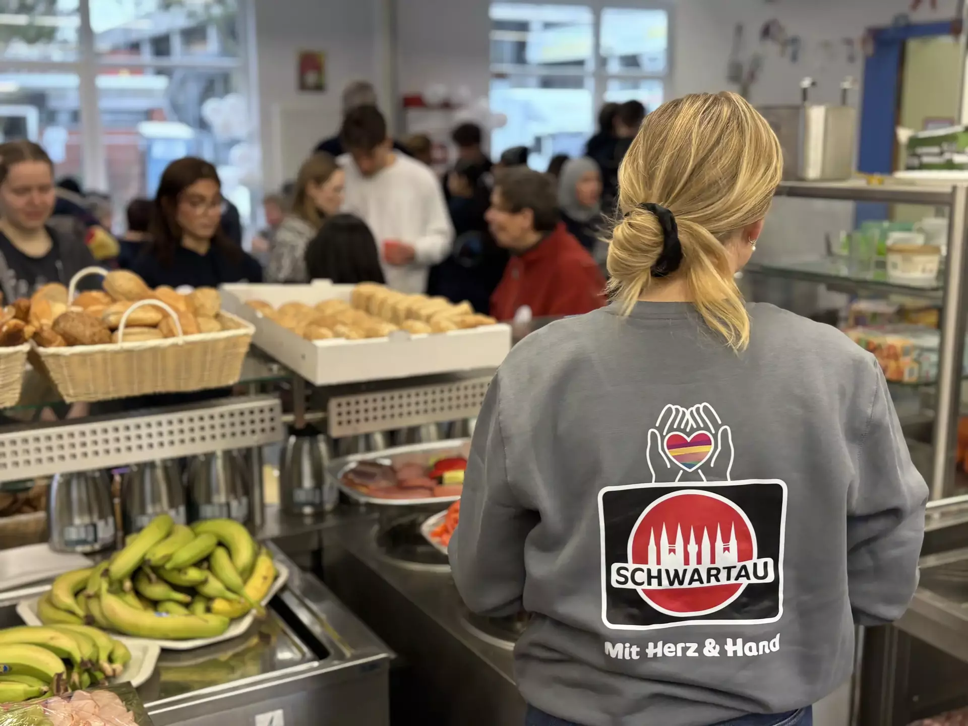 Man sieht den Rücken eine Kollegin mit dem großen Schwartau-Logo bedruckt am Ausgabe-Buffett, vorne Körbe mit Obst und Brötchen.