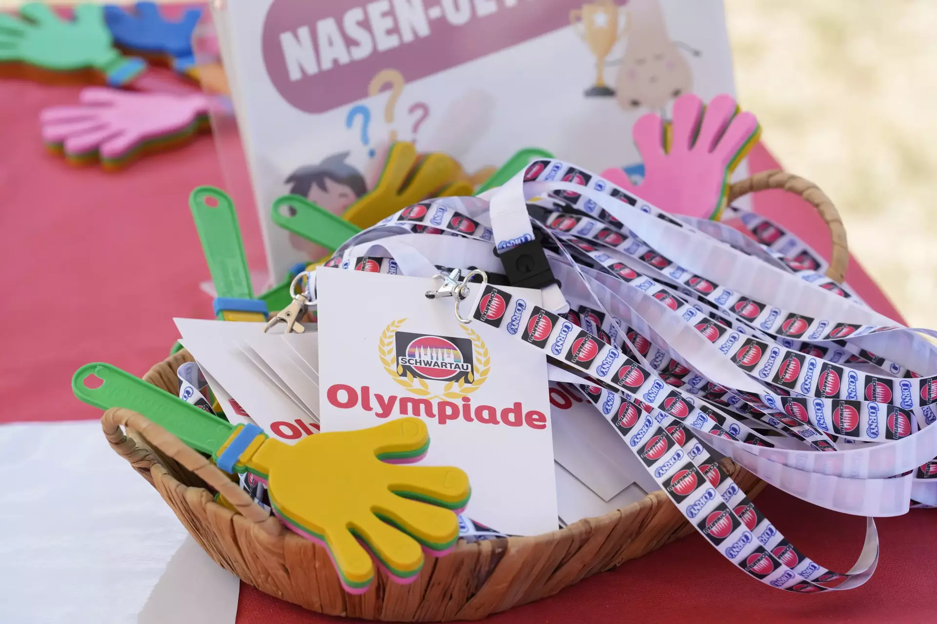 Auf dem Bild ist ein bunter und einladender Tischaufbau zu sehen, der zur Schwartau-Olympiade bei den Special Olympics gehört. Im Mittelpunkt steht ein geflochtener Korb, der mit Lanyards (Schlüsselbändern) mit dem Schwartau-Logo und großen Namenskarten mit der Aufschrift „Olympiade“ gefüllt ist. Die Lanyards sind weiß mit Schwartau-Logos in einer Reihe bedruckt.  Außerdem befinden sich im Korb und drumherum bunte Klatschhände aus Schaumstoff und ein Aufsteller im Hintergrund mit der Aufschrift „NASEN-OLYMPIADE“, begleitet von lustigen Zeichnungen, z. B. einer Nase mit Pokal. Das Ganze wirkt spielerisch, freundlich und ist offensichtlich auf Kinder oder Jugendliche zugeschnitten – perfekt passend zur fröhlichen, inklusiven Atmosphäre der Special Olympics.  Die Szene strahlt Spaß, Teamgeist und Engagement aus – mit viel Liebe zum Detail gestaltet.