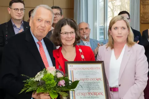 Die feierliche Ehrung von Dr. Arend Oetker als Ehrenbürger Bad Schwartaus im Sitzungssaal des Rathauses mit Holzverkleidung und großen Fenstern. Im Mittelpunkt steht Dr. Arend Oetker im Anzug mit roter Krawatte, der die Ehrenurkunde als 4. Ehrenbürger hält und einen Blumenstrauß trägt. Neben ihm stehen die Bürgermeisterin Dr. Katrin Engeln und die Stadtpräsidentin Wiebke Zweig, eine in einem roten Blazer, die ebenfalls die Urkunde hält, und eine in einem rosafarbenen Hosenanzug. Im Hintergrund applaudieren mehrere festlich gekleidete Personen.