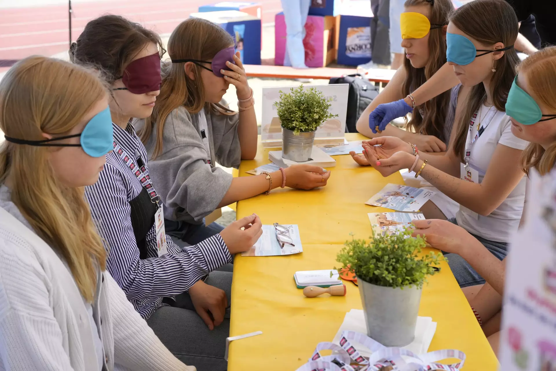 Auf dem Bild sieht man eine Gruppe junger Menschen, die an einem gelben Tisch sitzen – alle mit bunten Augenbinden. Es handelt sich um eine Station der Schwartau-Olympiade, vermutlich den „Schmeckathlon“, bei dem eine Blindverkostung stattfindet. Eine Person mit blauer Einweghandschuhen reicht gerade ein Produkt zum Probieren. Vor den Teilnehmenden liegen kleine Flyer, vermutlich zur Orientierung oder für Notizen.  Die Szene zeigt konzentrierte Gesichter und eine ruhige, neugierige Atmosphäre. Durch die Augenbinden stehen Geschmack und Sinne im Mittelpunkt – ein kreativer Ansatz, um Barrieren spielerisch zu thematisieren und Erlebnisse zu schaffen, die verbinden.  Im Vordergrund liegen wieder Schwartau-Lanyards, außerdem Topfpflanzen als freundliche Deko – alles liebevoll gestaltet. Das Bild strahlt Gemeinschaft, Teilhabe und Inklusion aus.