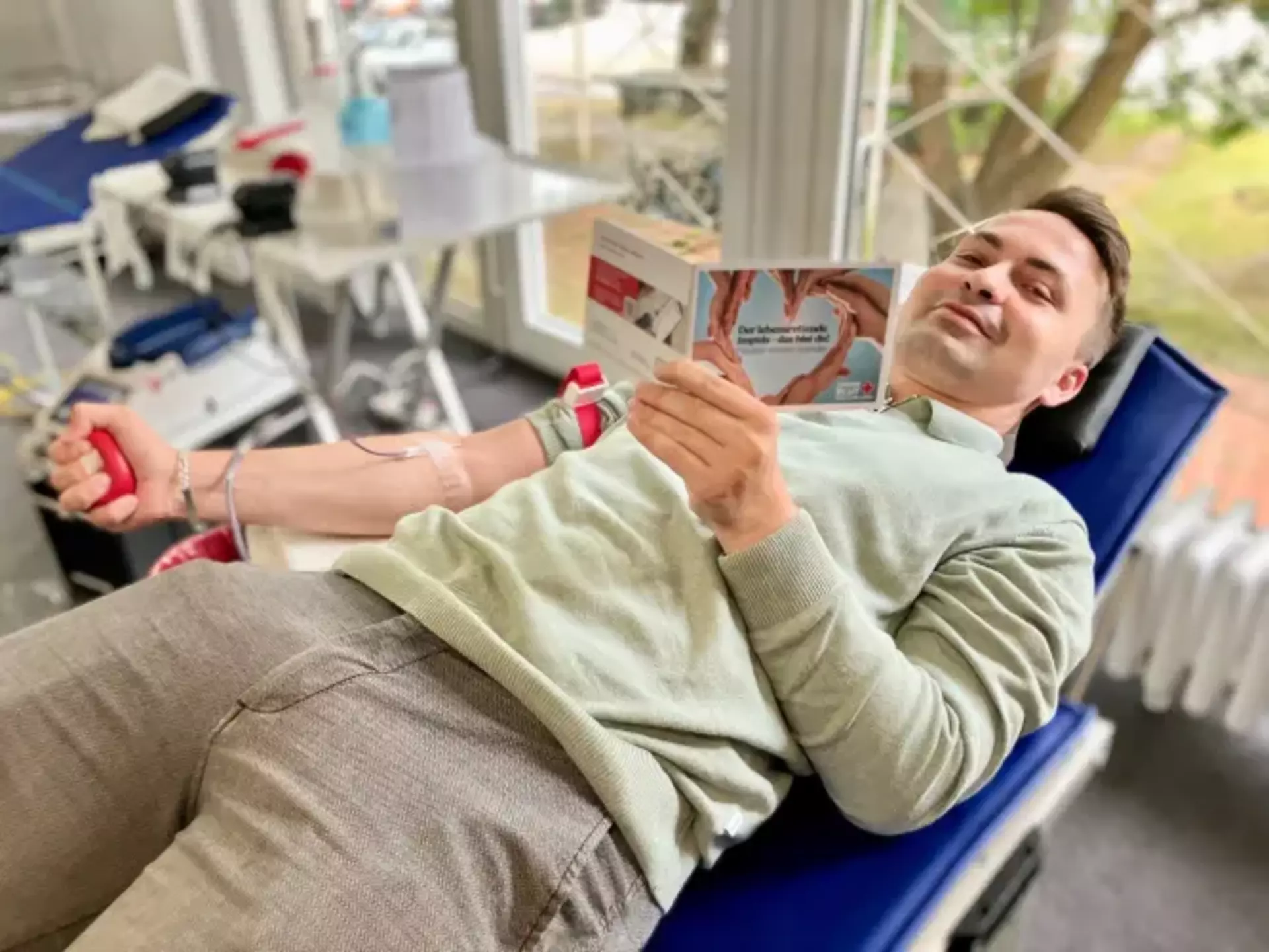 Hannes aus dem Marketing spendet Blut auf einer medizinischen Liege, hält ein Informationsheft und drückt mit der linken Hand einen roten Stressball – im Hintergrund medizinisches Equipment und große Fenster mit Tageslicht.