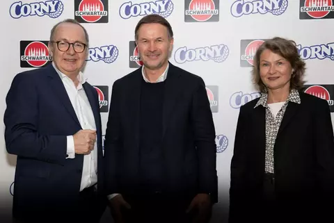 Drei Personen - Ralf Nagel, Markus Kohrs-Lichte und Heike Reimann - stehen vor einer Pressewand mit den Logos von „Schwartau“ und „Corny“. Sie tragen Business-Kleidung und lächeln in die Kamera. Links ein Mann mit Brille in einem dunklen Anzug, in der Mitte ein Mann mit dunklem Blazer und hellem Hemd, rechts eine Frau in einem dunklen Blazer mit gemusterter Bluse