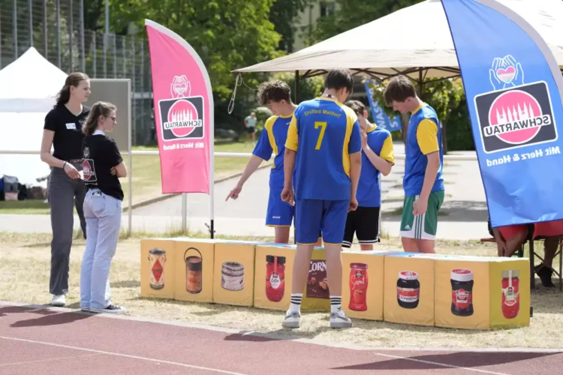 Auf dem Bild ist eine sportliche und fröhliche Atmosphäre zu sehen. Eine Gruppe junger Menschen in blauen Trikots mit gelben Akzenten (Team „Unified Meldorf“) steht vor einer Spielstation der Schwartau-Olympiade. Die Teilnehmenden zeigen auf die abgebildeten Produkte – es scheint sich um die Station „Würfel-Spaß“ zu handeln, bei der Schwartau-Produkte in die richtige Reihenfolge gebracht werden sollen.  Zwei Mitarbeiterinnen in schwarzen Schwartau-Shirts stehen betreuend daneben und begleiten die Aktion. Bunte Schwartau-Fahnen mit dem Slogan „Mit Herz & Hand“ rahmen das Bild ein und unterstreichen das Engagement des Unternehmens.  Die Szene vermittelt Teamgeist, Aktivität und Spaß – kombiniert mit der klaren Botschaft: Inklusion leben, Gemeinschaft stärken.