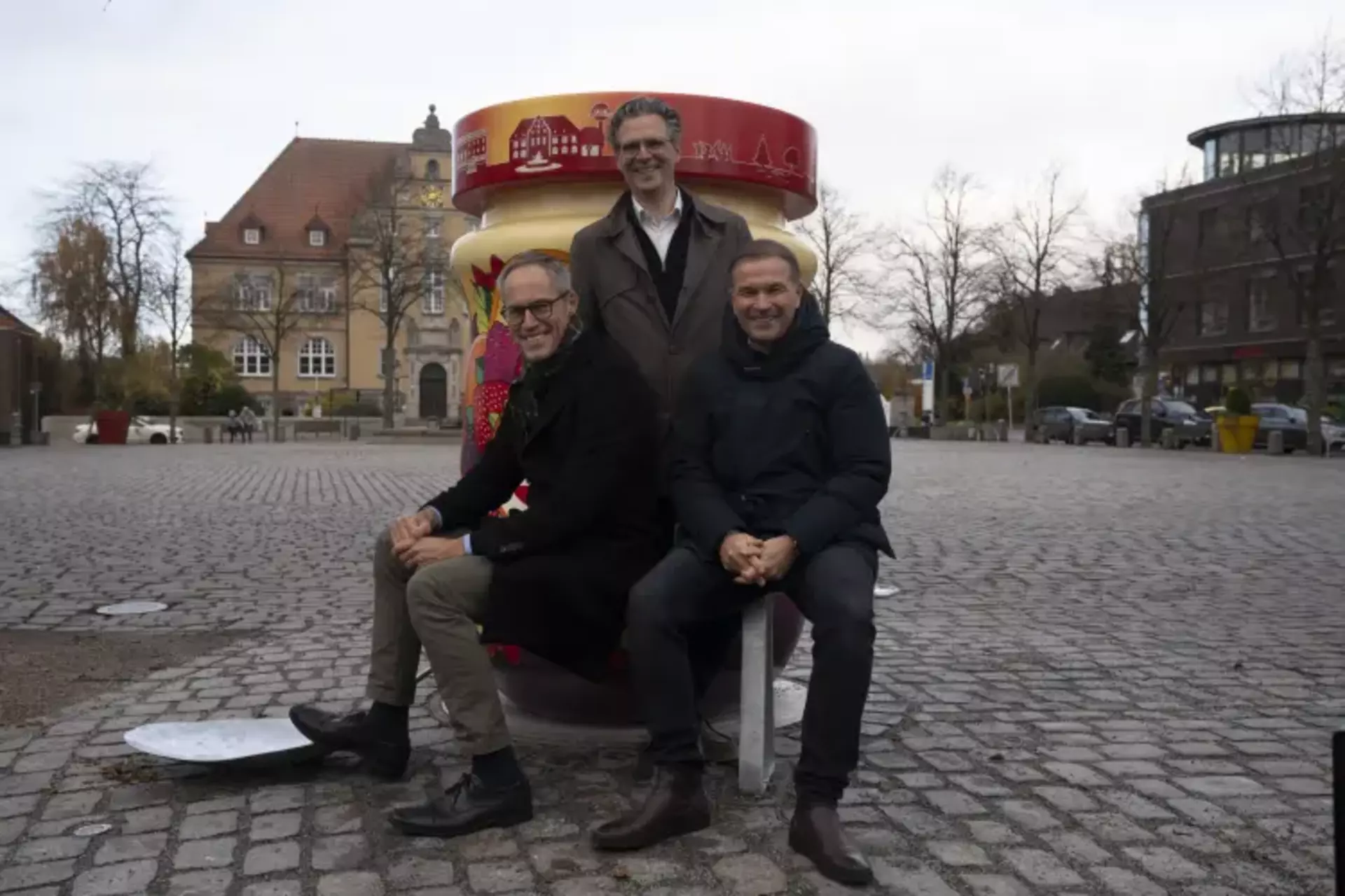 Auf dem Bild sieht man den den Fotopoint "Marmeladenstadt", im Hintergrund der Schwartauer Marktplatz und die drei Geschäftsführer der Schwartauer Werke auf einem Metalllöffel vor dem Fotopoint,