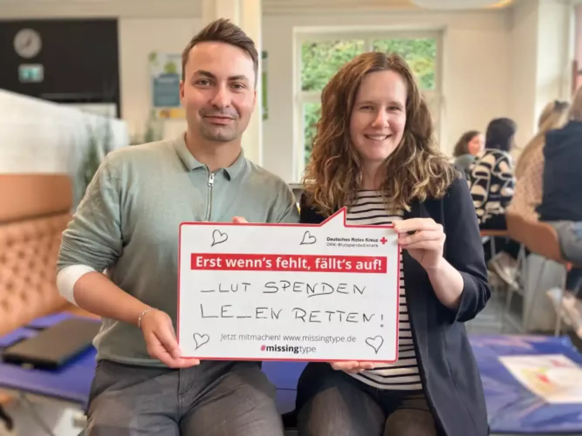 Hannes und Henrieke aus dem Marketing halten ein Schild mit der Aufschrift: ‚Erst wenn's fehlt, fällt's auf! _LUT SPENDEN LE_EN RETTEN! Jetzt mitmachen! www.missingtype.de #missingtype‘ – eine Aktion zur Blutspende, bei der bewusst Buchstaben weggelassen wurden, um auf ihre Bedeutung aufmerksam zu machen.