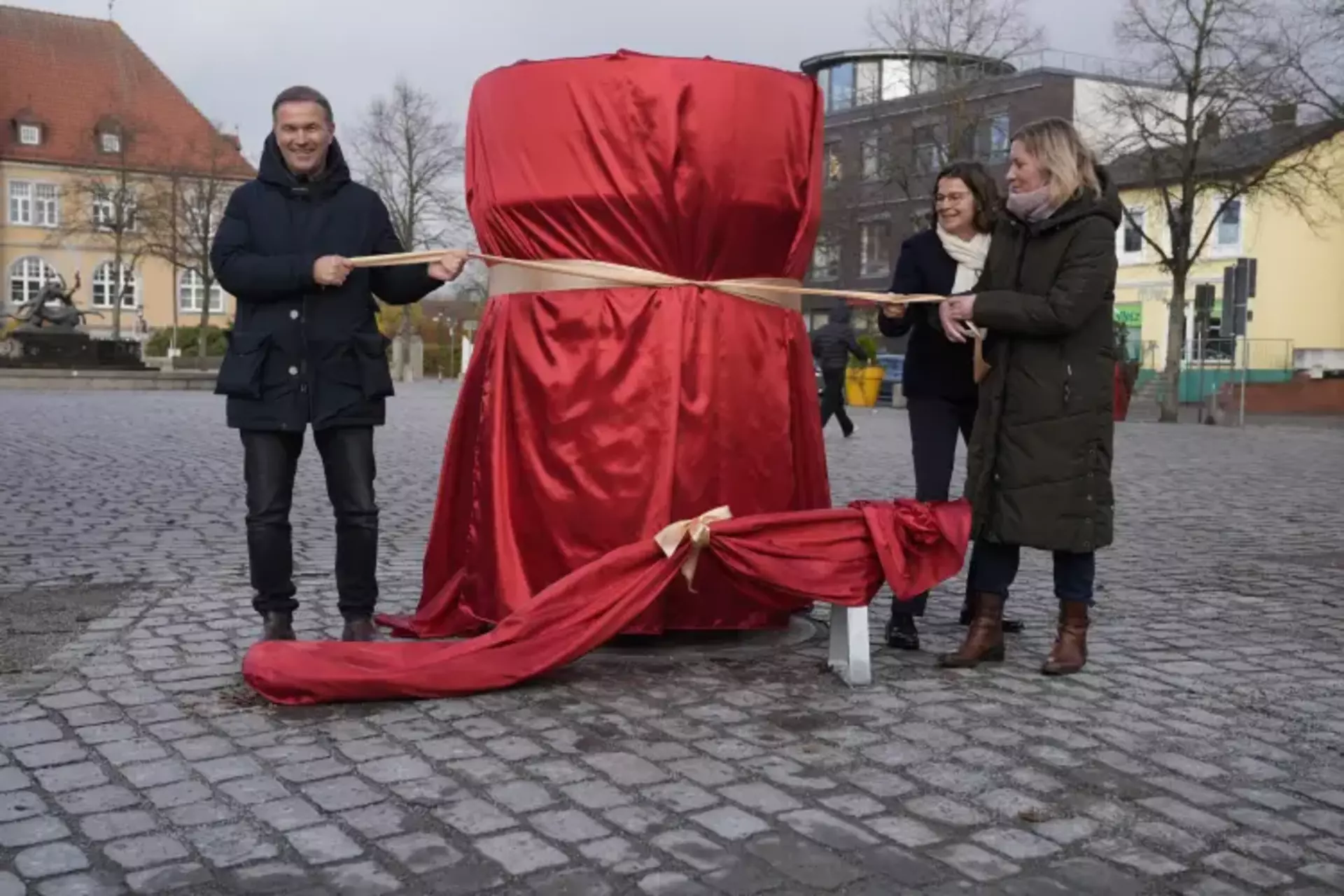 Aud dem Bild steht links ein Mann und rechts zwei Damen neben einem großen verfüllten Objekt in rot mit einer goldenen Schleife. Alle ziehen an der Schleife.