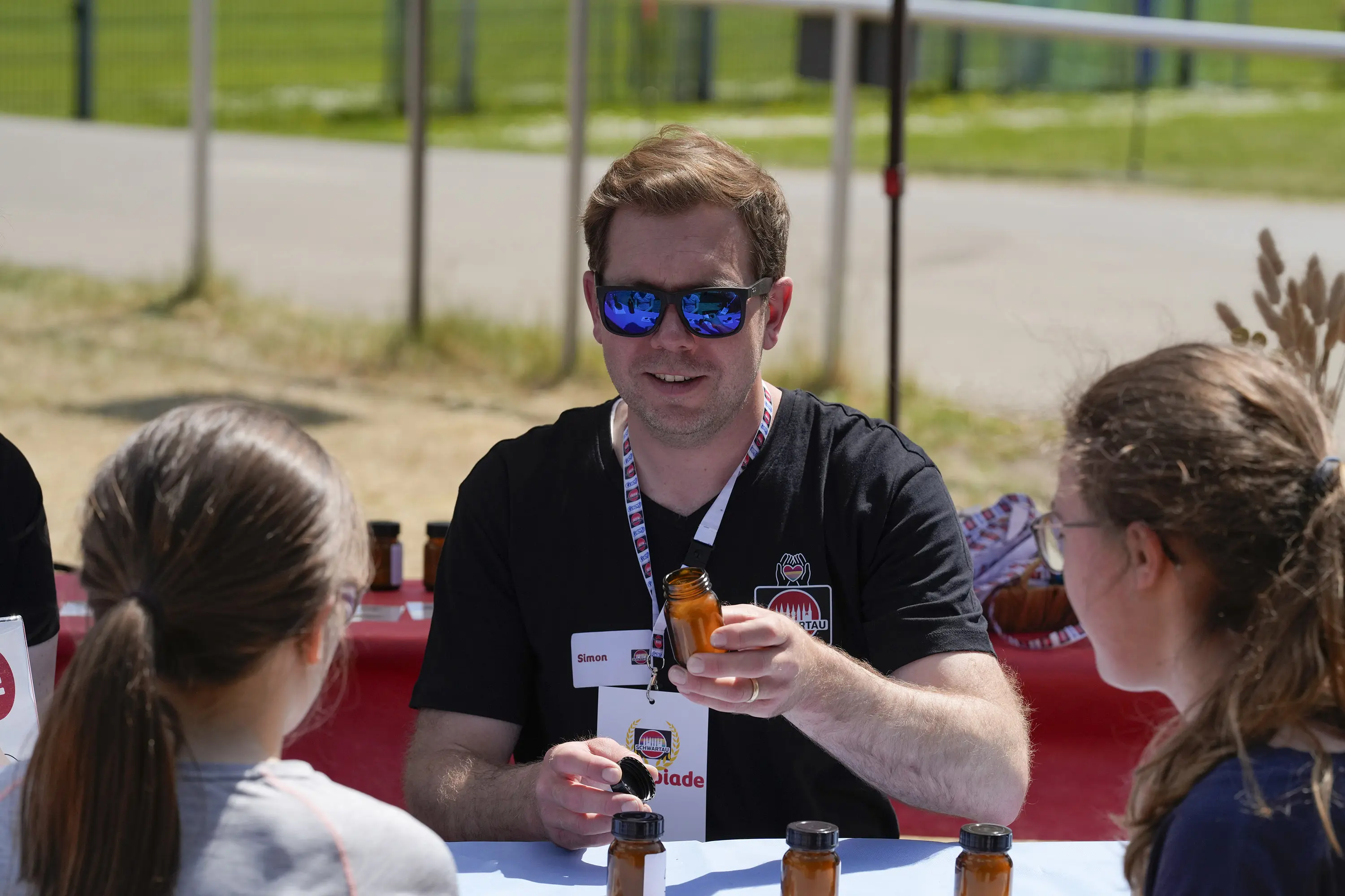 Auf dem Bild sieht man Simon Bruhn, Manager Factory Planning & Logistics, der auf einem Sportplatz auf einer Bierzeltgarnitur sitzt. Vor ihm - mit dem Rücken zum Bild - sitzen zwei junge Mädchen, die ihn interessiert anschauen. Simon hält eine Flasche mit einer Duftprobe hoch, er begleitet die Nasenolympia, bei der man verschiedene Aromen erreichen muss. 