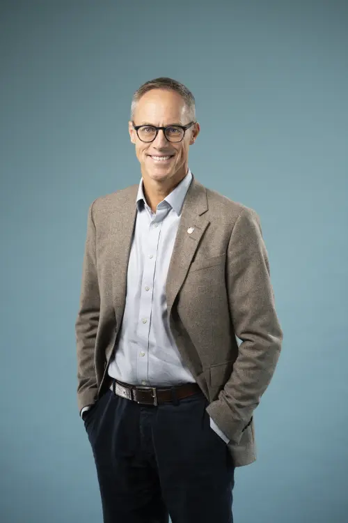 Ein schlanker Mann mit Brille und grauen kurzen Haaren, mit hellem Sacko, dunkler Jeans und Hemd vor einer Leinwand. Freundliches Lächeln