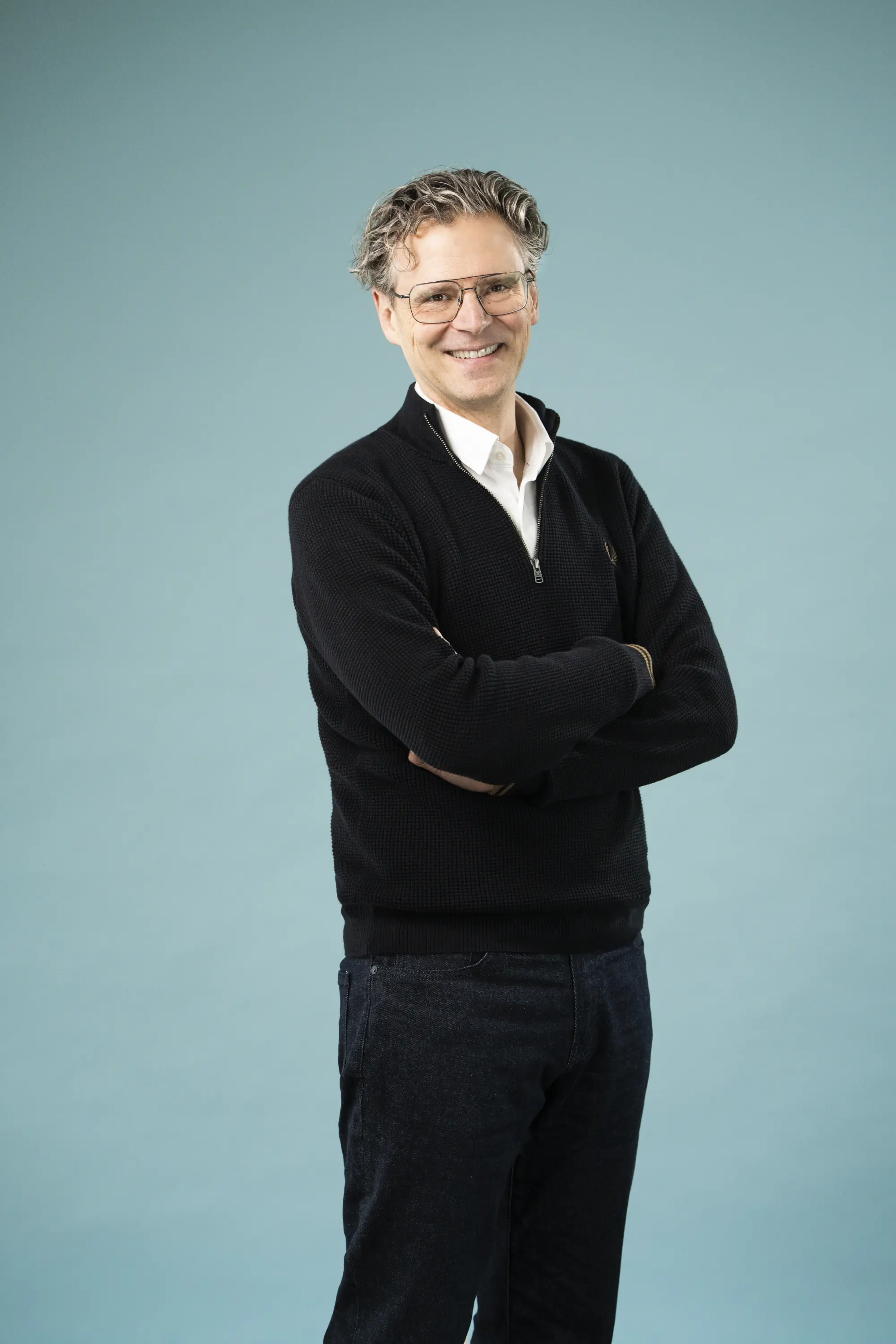 Schlanker Mann mit Brille, dunkle Hose & Pullover vor einer Leinwand, freundliches Lächeln