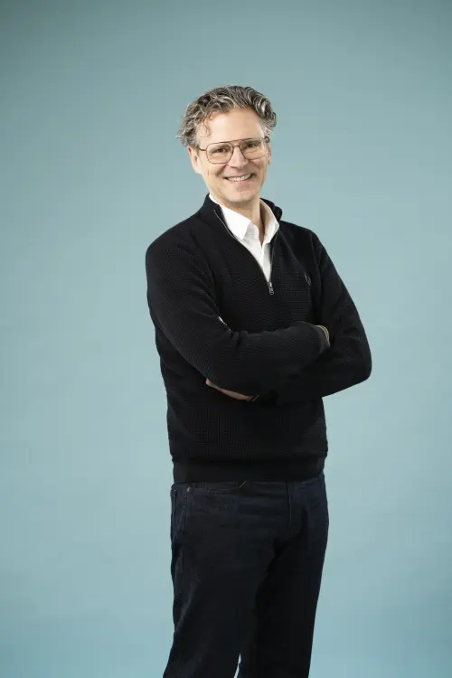 Schlanker Mann mit Brille, dunkle Hose & Pullover vor einer Leinwand, freundliches Lächeln