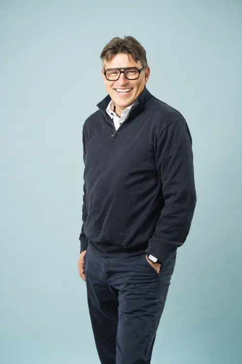 Schlanker Mann mit Brille, dunkle Hose & Pullover vor einer Leinwand, freundliches Lächeln