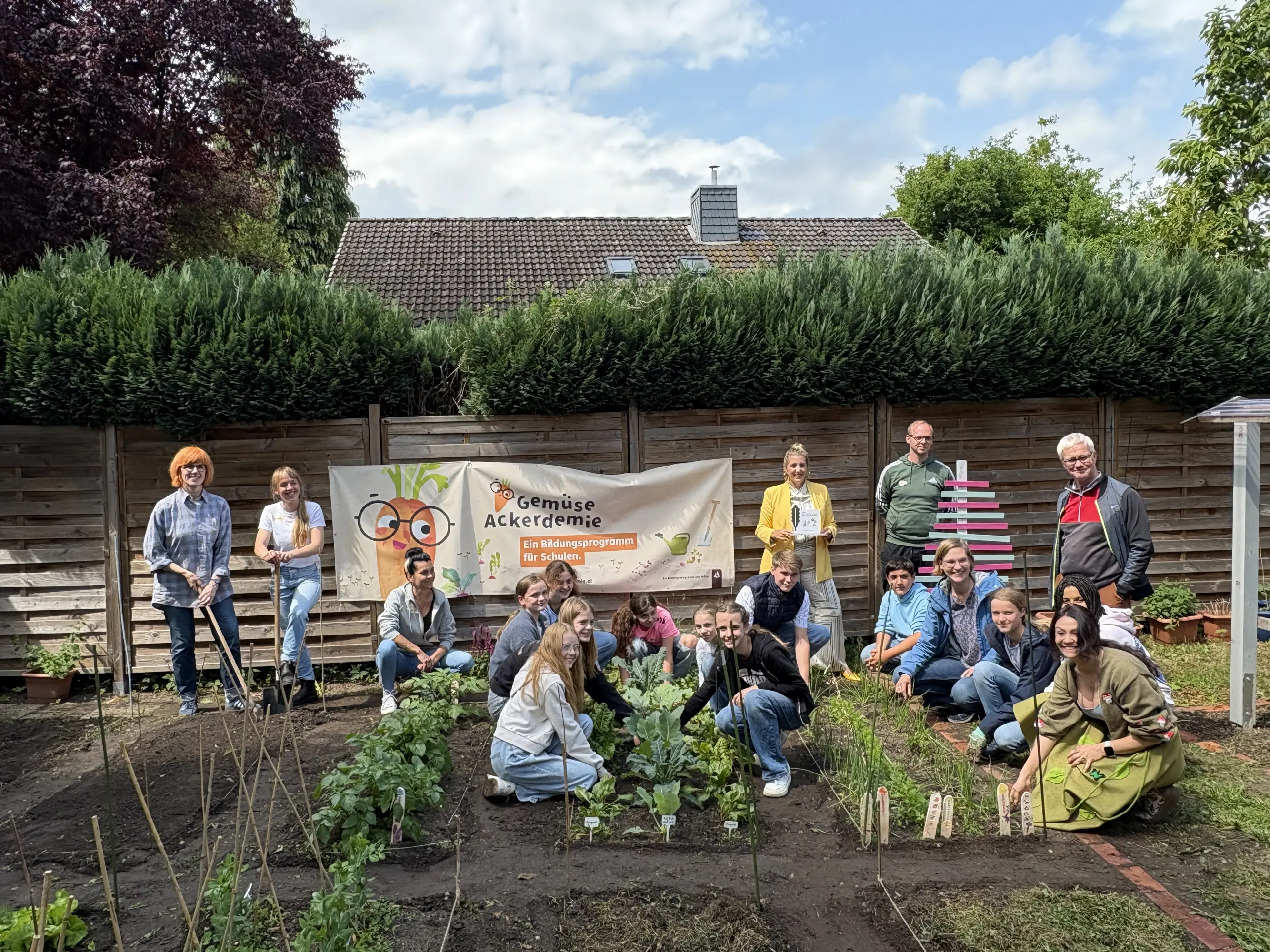 Auf dem Foto sind eine Gruppe von Schüler*innen der Klasse 6b der Elisabeth-Selbert-Gemeinschaftsschule, AckerCoach Nicola Sanftleben-Pychynski, Maike Ladendorf (Lehrerin), Andrea Linke (Mitarbeiterin Offene Ganztagsschule), die AckerKinder aus der 6. Klasse, Vanessa Krabiell (Schwartauer Werke), Nino Dubanowski (Leiter Offene Ganztagsschule), Michael Thole (Bürgerverein Bad Schwartau) und Stephanie Thieß (Schulleitung) zu sehen. Sie stehen oder hocken rund um mehrere Beete mit verschiedenen Gemüsepflanzen, die gepflegt und beschriftet sind. Im Hintergrund hängt ein Banner mit der Aufschrift „GemüseAckerdemie – Ein Bildungsprogramm für Schulen“, das auf das Bildungsprojekt hinweist. Einige Personen halten Gartenwerkzeuge, andere lächeln in die Kamera. Die Stimmung ist fröhlich und gemeinschaftlich. Der Garten ist gut gepflegt, mit klaren Beeten und einer dekorativen, pink-weißen Holzinstallation. Der Ort ist wie ein grüner Lern- und Begegnungsraum  auf Gelände der Schule. Das Foto vermittelt Engagement für nachhaltige Bildung und Umweltbewusstsein.